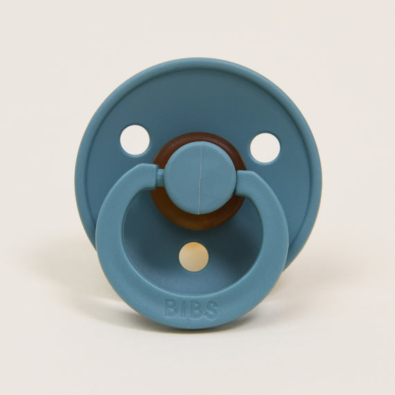 Displayed against a plain white background is the Bibs Pacifier, part of the 2 Pack Island Sea collection. This pacifier features a blue color with a round shield and three ventilation holes, along with a brown center and handle. It is crafted from natural rubber and designed and manufactured in Denmark, with "BIBS" embossed on the handle to help soothe little ones.