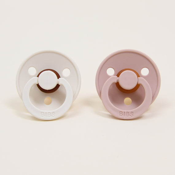 The Bibs Pacifier Set, featuring two round baby pacifiers with a classic design, is displayed against a plain background. The ivory natural rubber pacifier on the left contrasts with the blush self-soothe option on the right. Both pacifiers include a circular guard and handle.