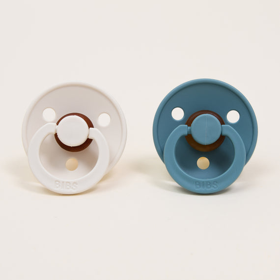 The Bibs Pacifier Set, featuring two round pacifiers made from natural rubber, is shown against a neutral background. The pacifier on the left is Ivory, and the one on the right is Island Sea. Designed in Denmark, both pacifiers include a circular handle and small ventilation holes on the shield to support soothing for your baby.