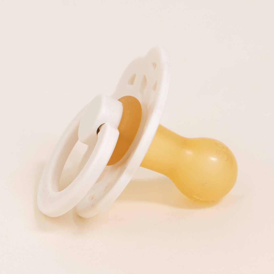 The Chloe Pacifier Set in Ivory & Vanilla showcases a natural rubber baby pacifier with a white shield and handle, complemented by a yellow nipple, all arranged on a plain, light-colored surface. Its bohemian-inspired design aids little ones in self-soothing.