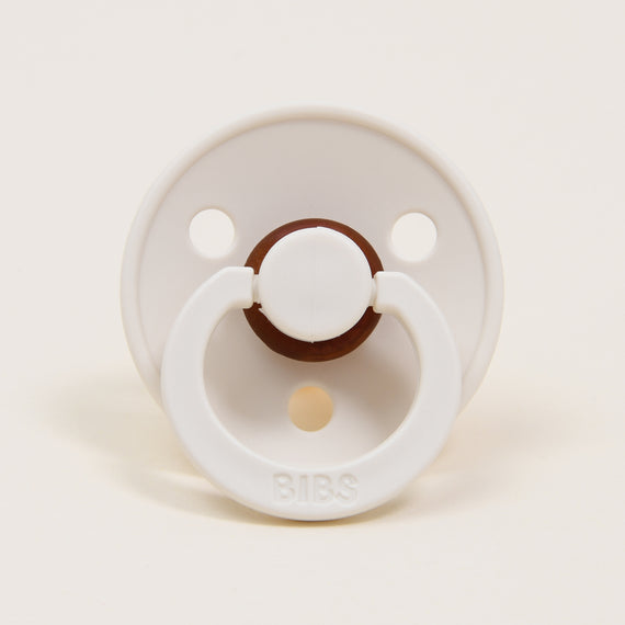 Among the showcased items, one is a Bibs Pacifier, known for its timeless elegance and crafted from natural rubber. It is displayed against a simple white backdrop, highlighting its ivory circular shield with two small openings at the top. The ring handle, embossed with "BIBS," is specifically designed to assist babies in self-soothing.