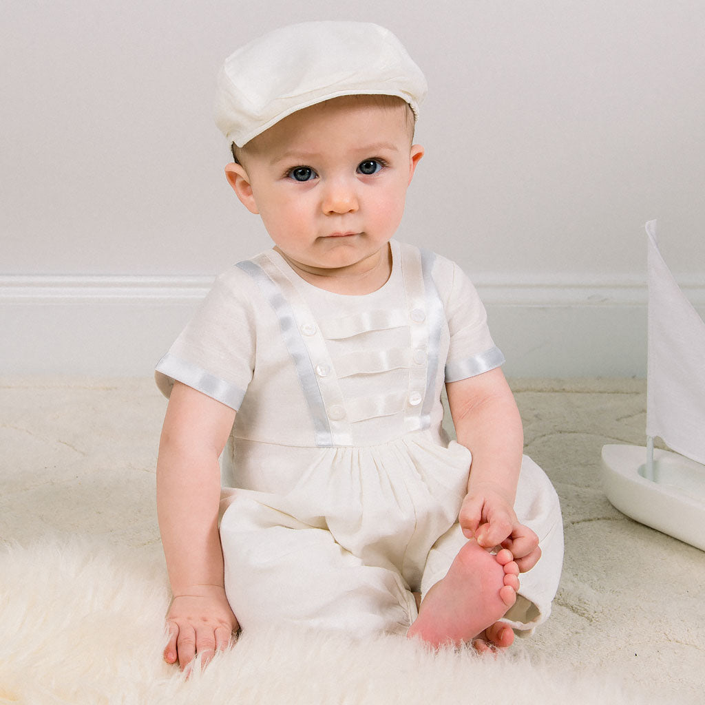 Baby boy sitting down and wearing the Owen Newsboy Cap and matching Linen Romper featuring silk ribbon in ivory and light blue across the front bodice and sleeves