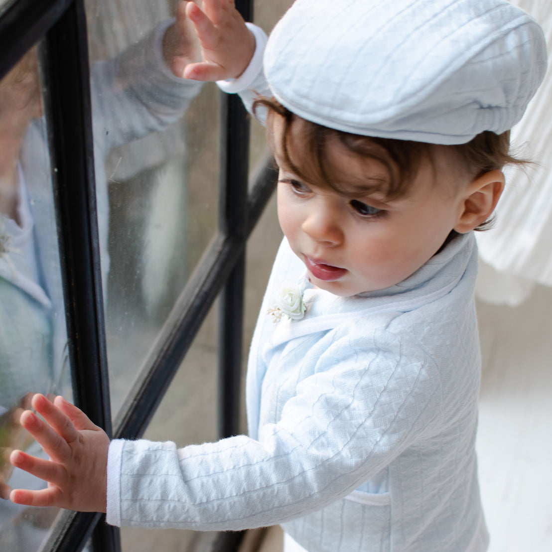 Baby boy looking outside the window and wearing the Logan 3-Piece Suit (and matching Newsboy Cap), including the jacket, pants and onesie. Jacket made from blue textured cotton with a white French terry cotton trim.