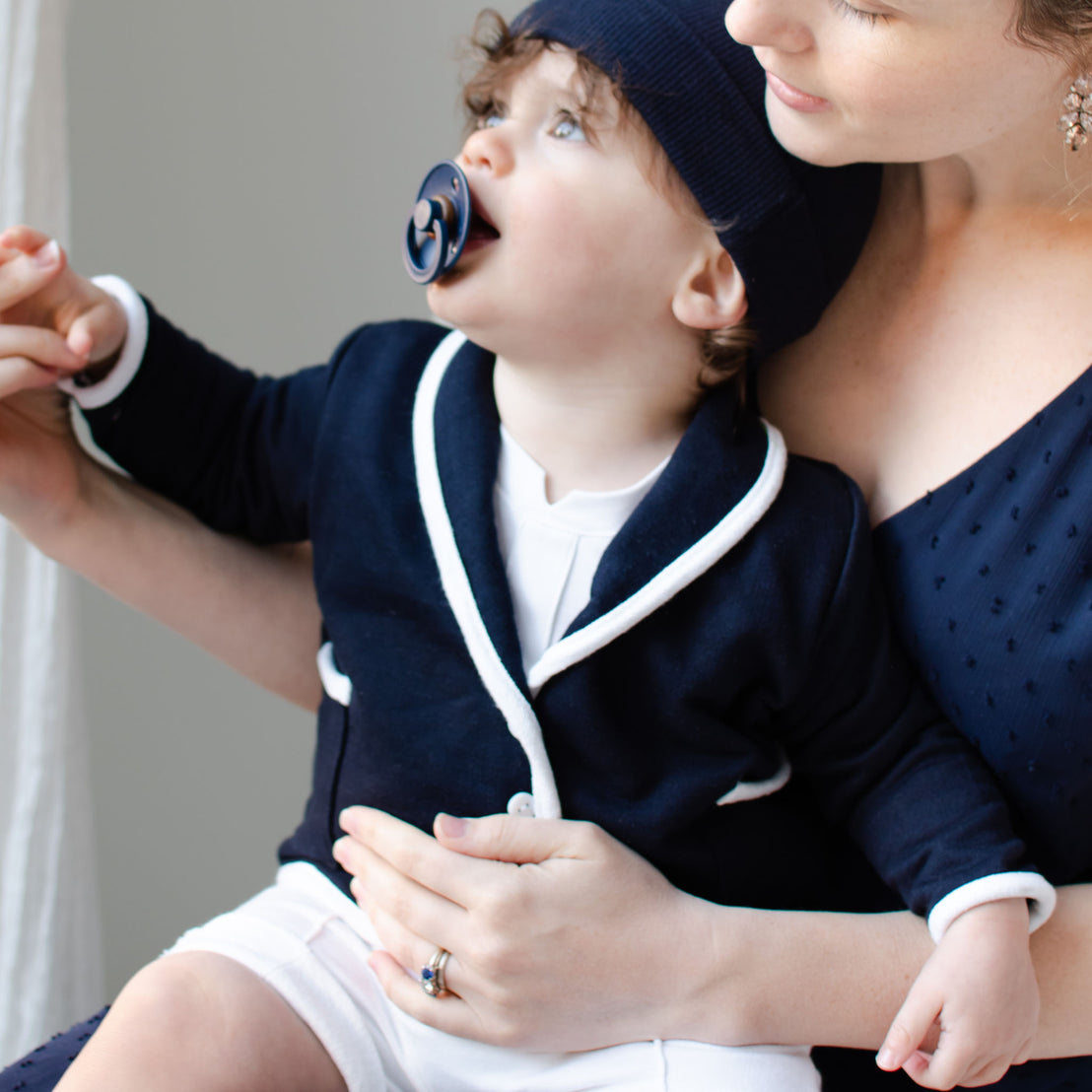 Baby boy sitting with his mother. He is wearing the Elliott 3-Piece Shorts Suit (with matching pacifier and Navy Ribbed Pima Beanie).