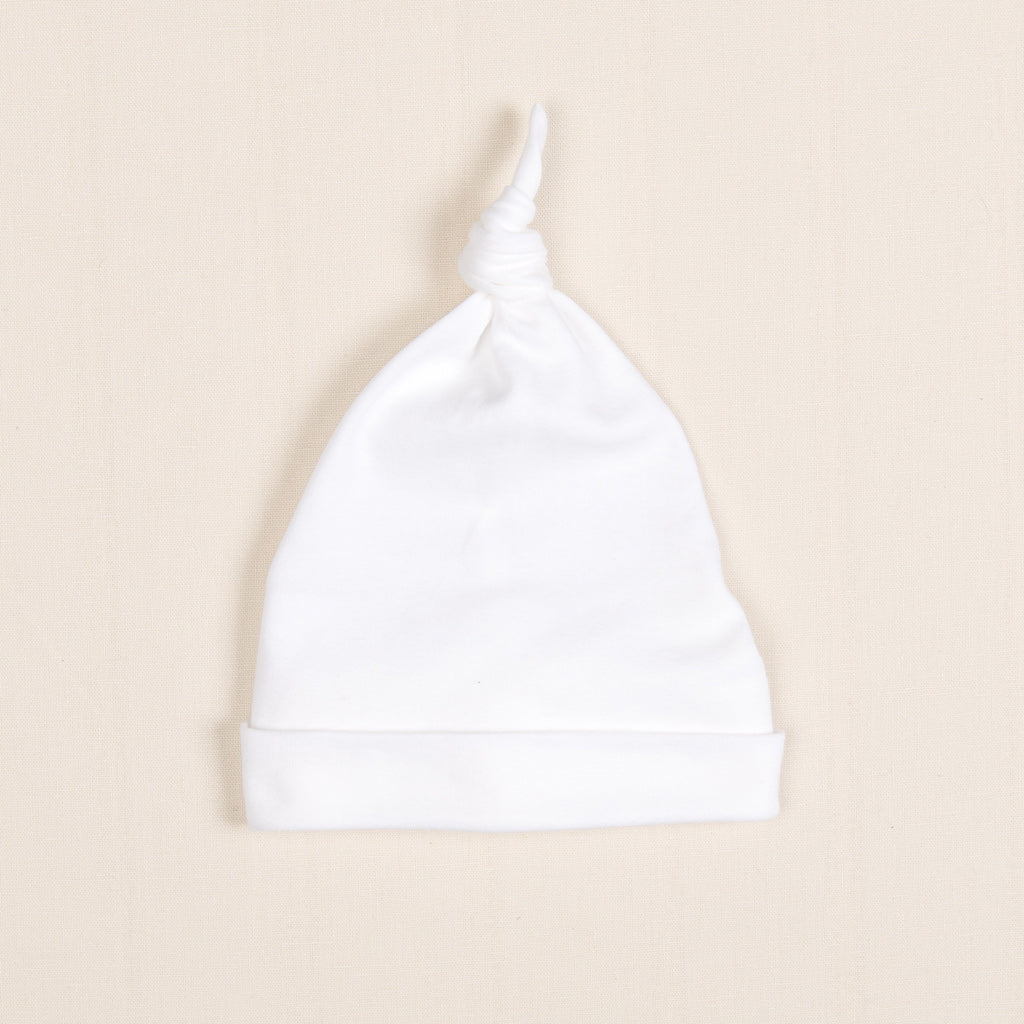The Newborn Knotted Cap | Liam, a small white hat crafted from luxurious pima cotton, rests on a light beige background.