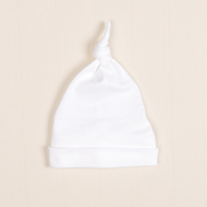 The Newborn Knotted Cap | Liam, a small white hat crafted from luxurious pima cotton, rests on a light beige background.
