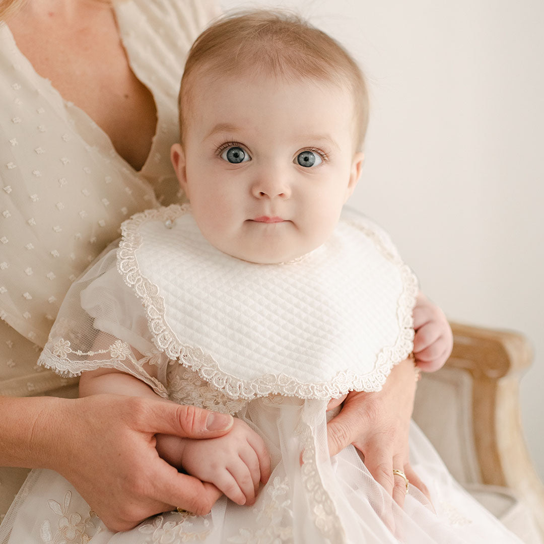 A baby with large eyes, wearing a delicate christening gown adorned with Venice lace and the Kristina Bib, is being held by an adult. The adult is in a light-colored dress, and both are seated against a neutral background.