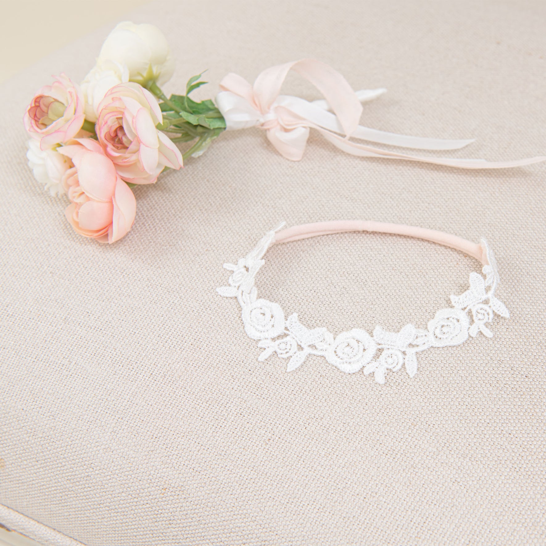 A Rose Lace Headband, adorned with delicate handcrafted lace roses, is displayed on a beige textured surface. Beside it rests a small bouquet of pink and white flowers tied with a ribbon, all softly illuminated to highlight their intricate features.