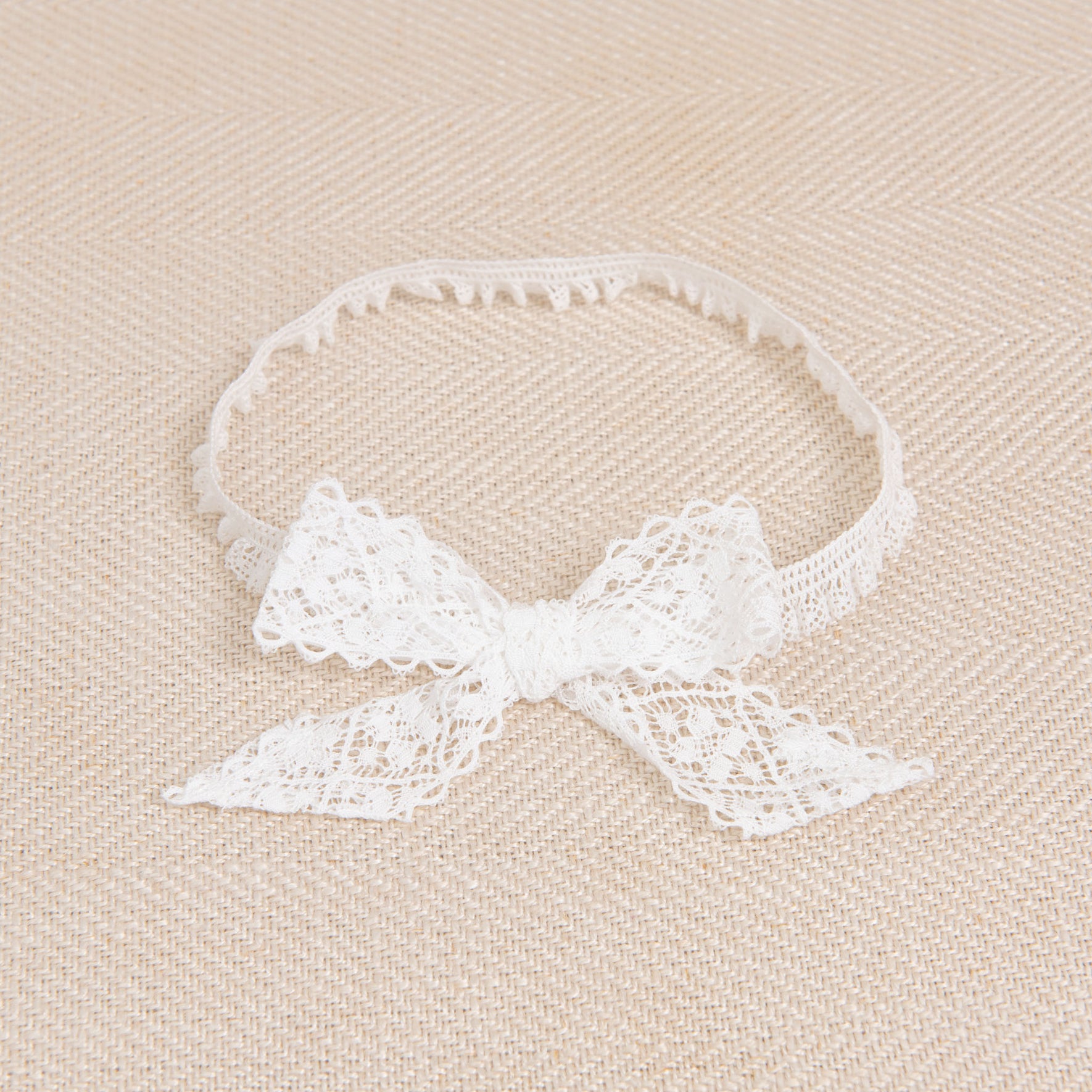 The Thea Lace Headband, handcrafted in the USA, is beautifully displayed on a beige textured surface. Its stretch lace features a delicate, intricate pattern, and the bow is neatly tied at the front.
