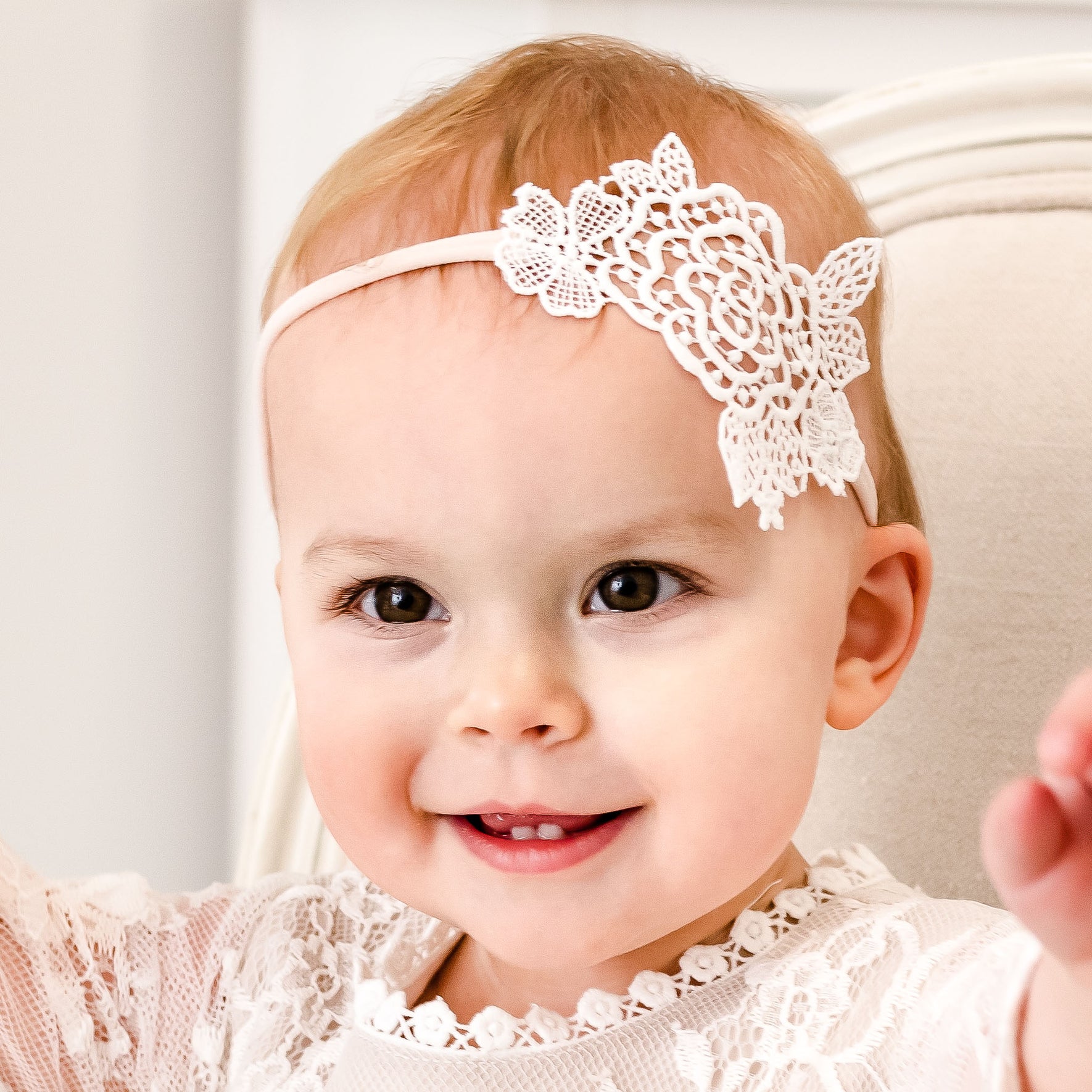A baby with light skin and short, light-colored hair is smiling. The baby is adorned in the Juliette Lace Headband featuring a decorative flower on the left side and dressed in an elegant light ivory rose lace outfit, handmade in the USA. They are sitting on a light-colored chair, radiating pure joy.