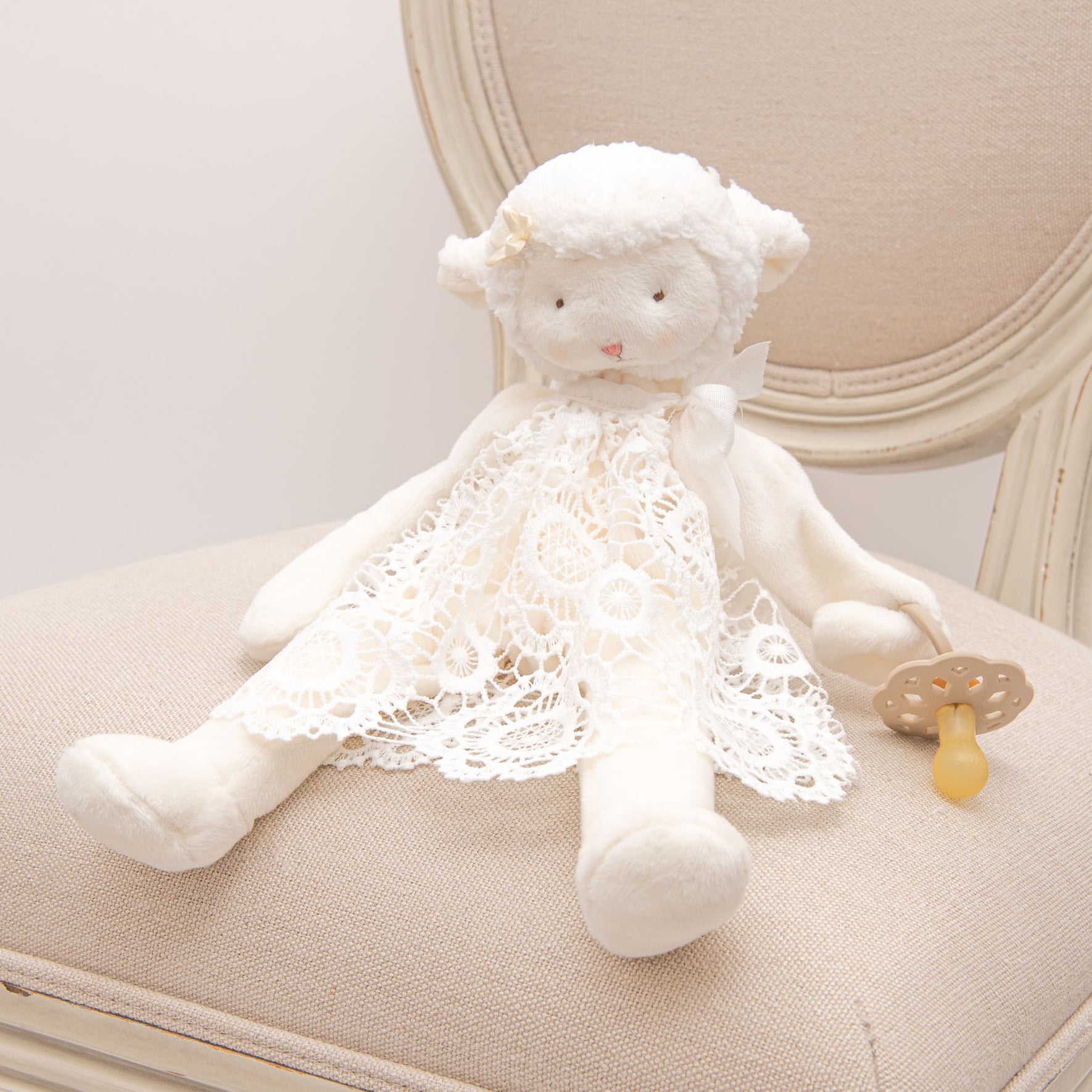 The Poppy Silly Lamb Buddy Pacifier Holder displayed on a beige chair, a soft lamb adorned with lace details coordinating with the Poppy Christening Gown, holding a pacifier.
