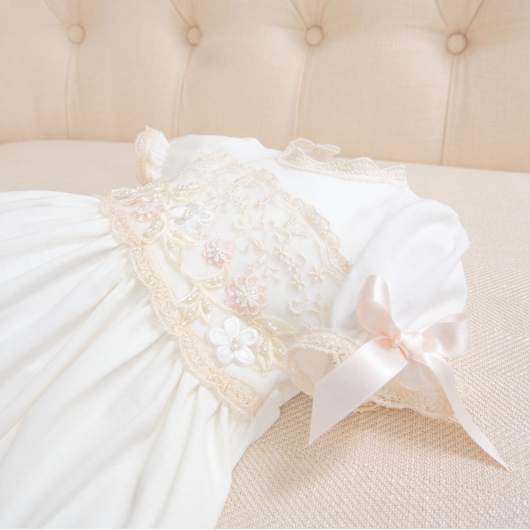 A close-up photograph of a Jessica Newborn Gift Set - Save 10% adorned with lace embroidery and small floral appliqués. The dress features short sleeves and a pink bow on one sleeve, perfect as a take me home outfit. It is placed on a beige, tufted background.