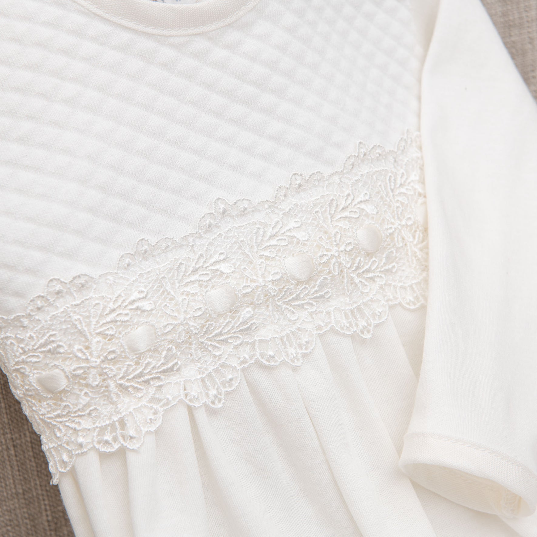 A close-up of the Madeline Newborn Gown crafted from soft pima cotton, featuring a quilted pattern on the upper section, delicate ivory lace trim with floral design and small fabric-covered buttons in the middle, and a pleated lower section. The sleeve on the right side is plain white.