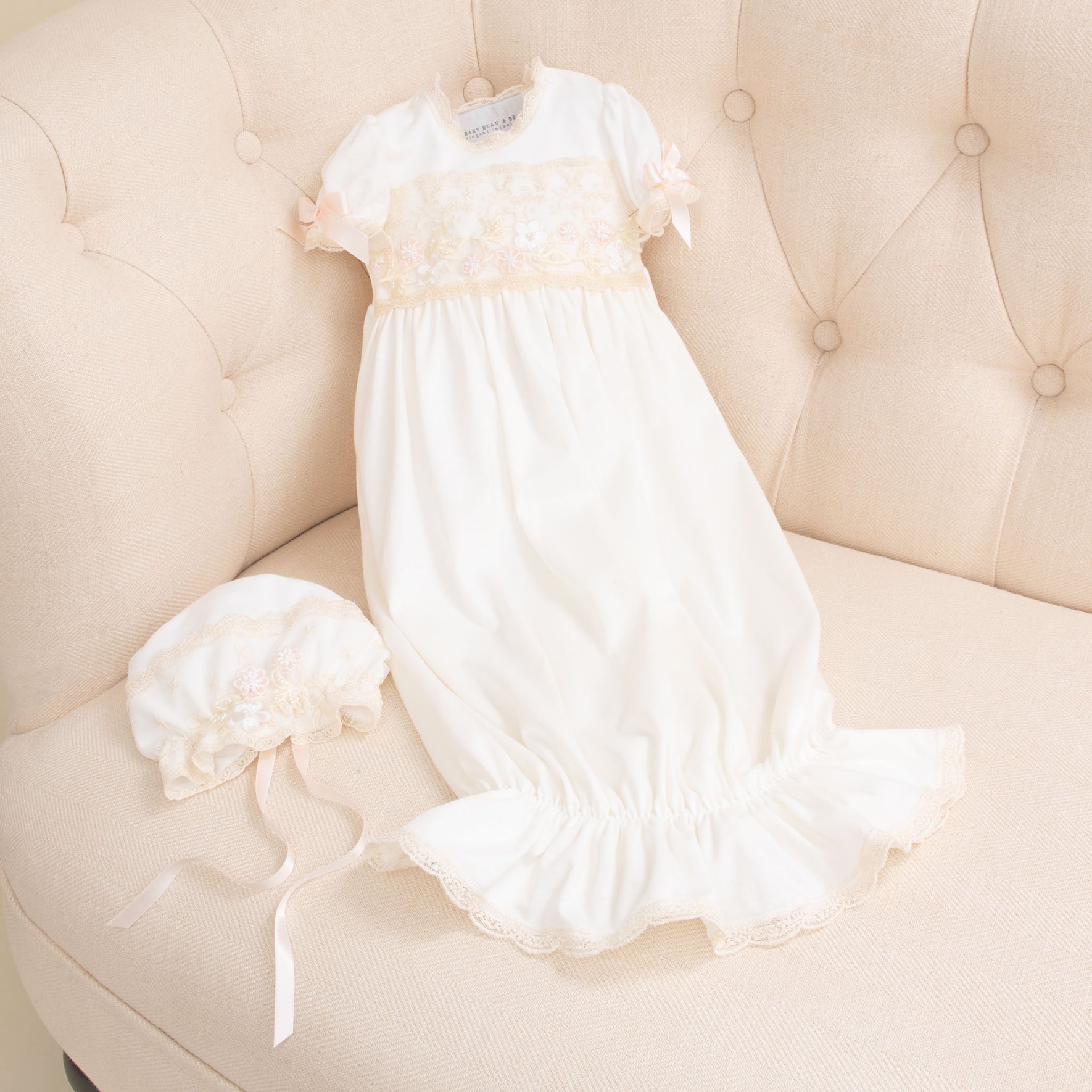 A Jessica Newborn Gift Set - Save 10% with lace detailing and a ruffled bottom is laid out on a cream-colored sofa. To the left of the set is a matching bonnet. Both items, handmade in the USA, feature delicate pink ribbon accents.
