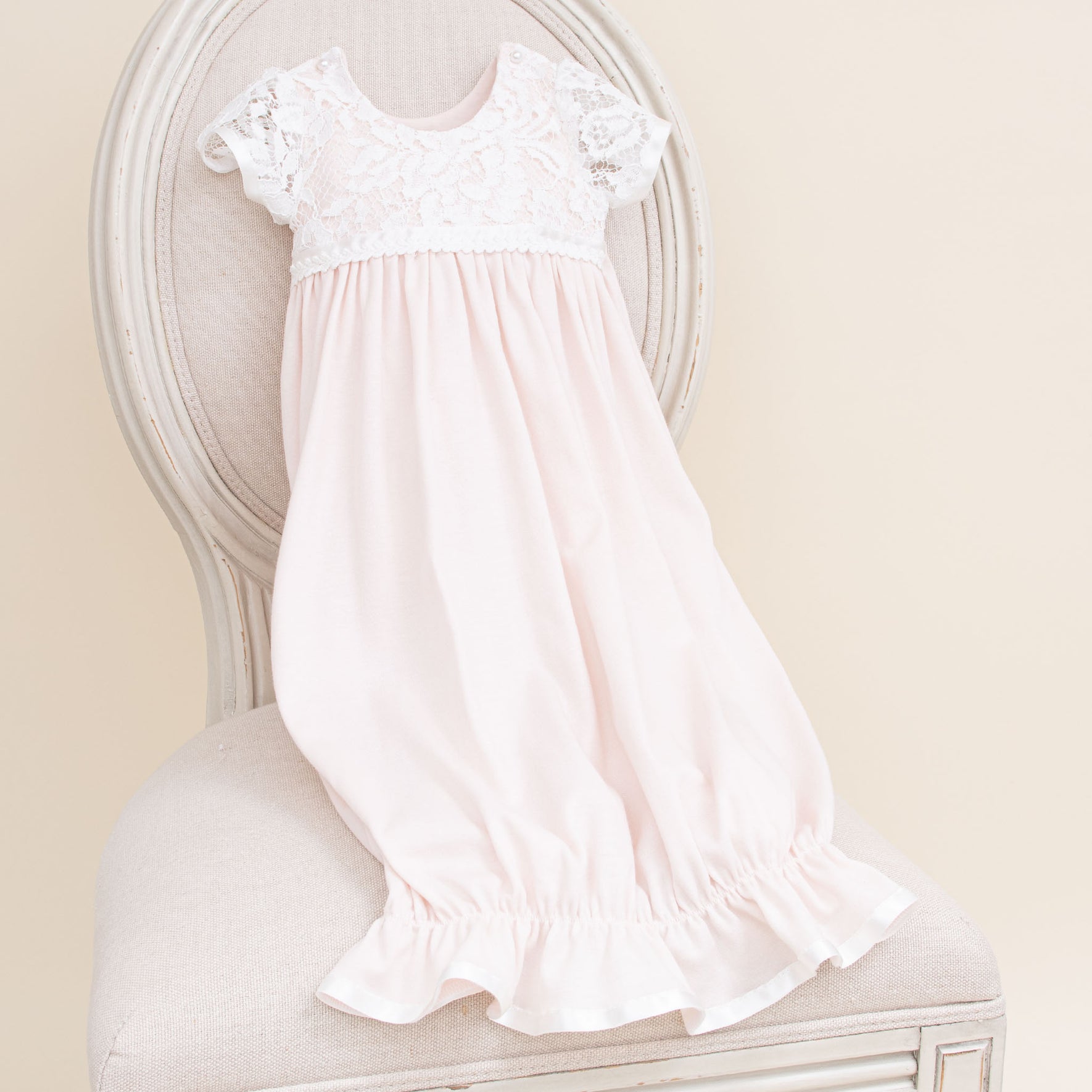 A blush cotton infant dress with short lace sleeves and a lace bodice is draped over a cushioned vintage-style chair. The dress has a gathered waist and ruffled hem, paired perfectly with the Rose Layette & Bonnet. The chair features light-colored upholstery with a rounded back and detailed wood trim.