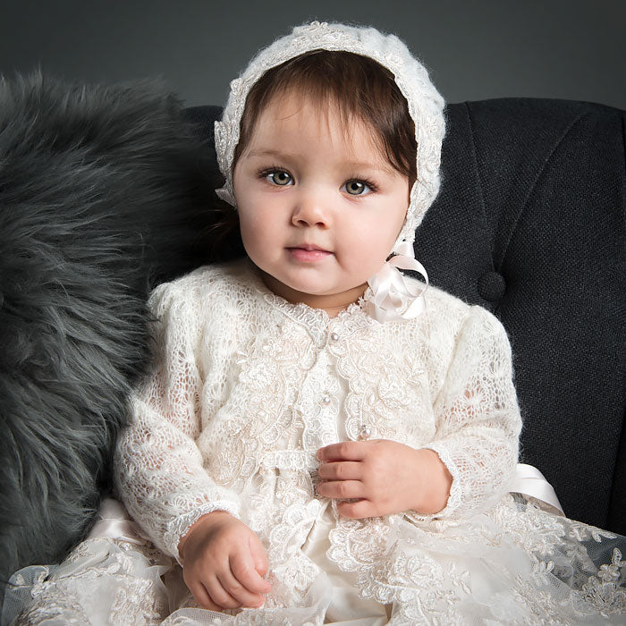 Baby girl looking into camera wearing her Penelope christening gown and knit sweater and bonnet.