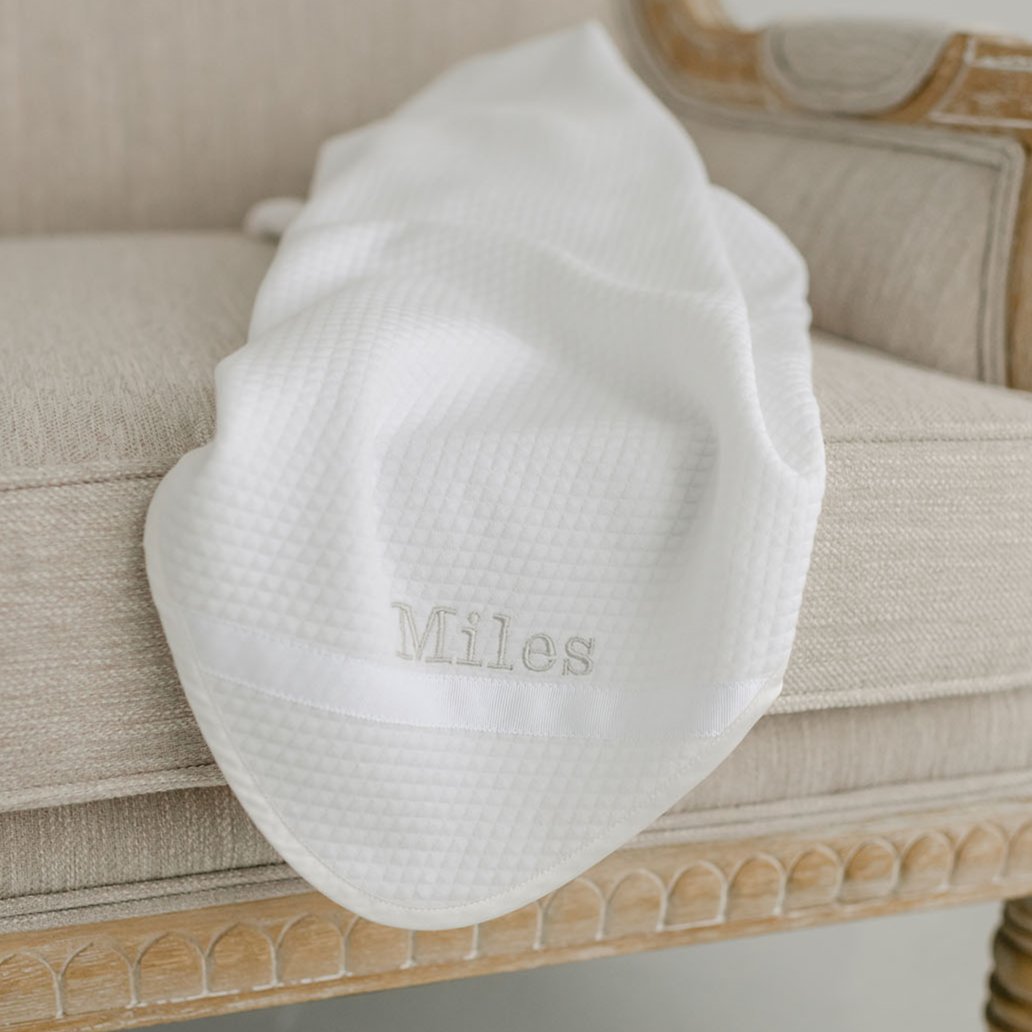 Flat lay photo of the Miles Personalized Blanket on a chair. It is made with quilted cotton and a ribbon detail on the corner. The name "Miles" is embroidered on the corner.