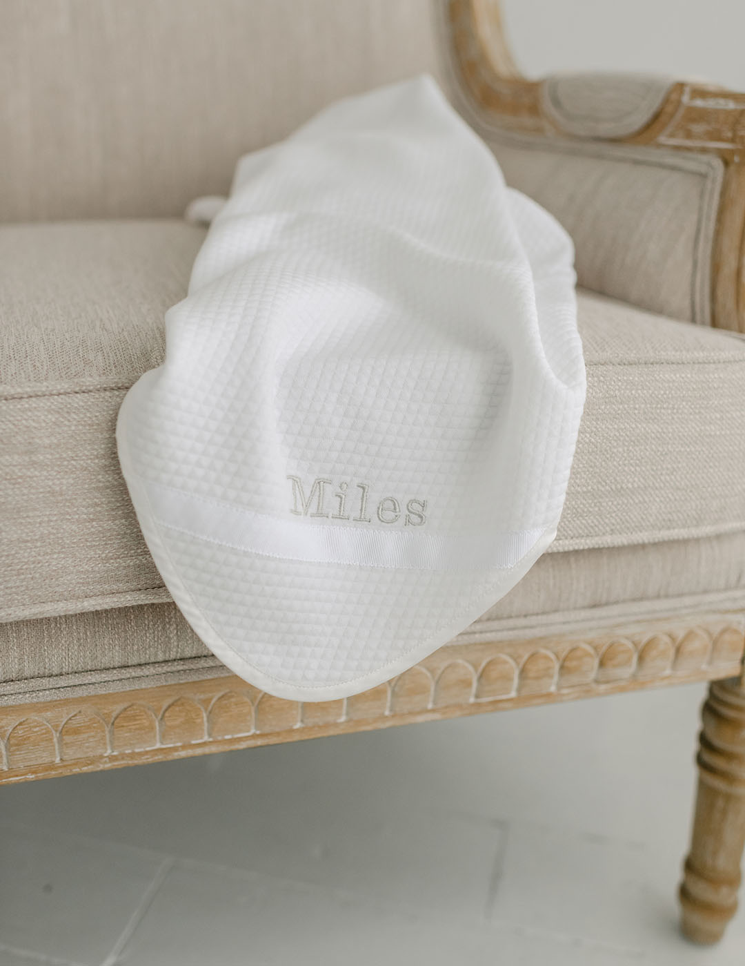 Flat lay photo of the Miles Personalized Blanket on a chair. It is made with quilted cotton and a ribbon detail on the corner. The name "Miles" is embroidered on the corner.