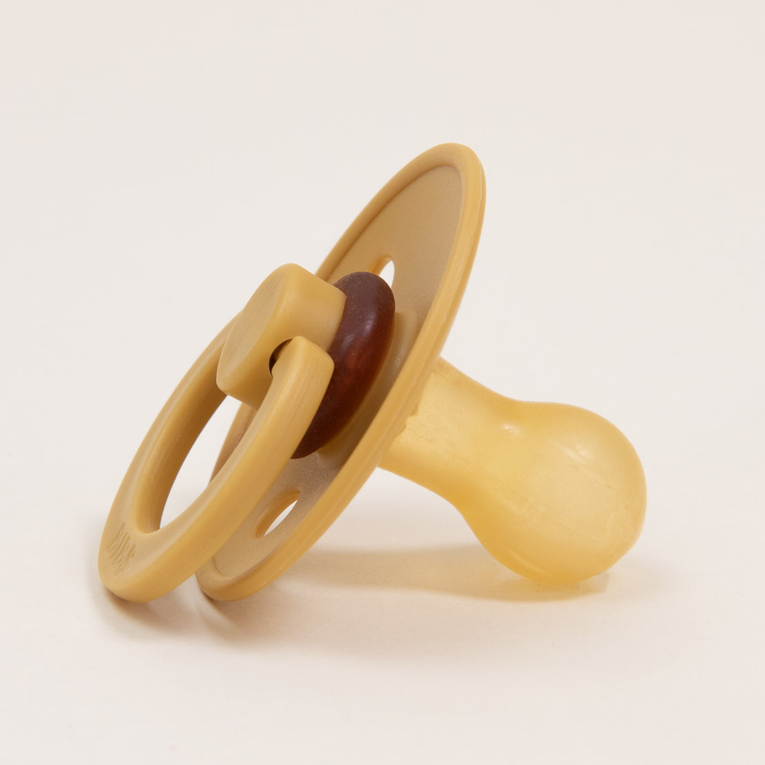 A mustard-colored Bibs Pacifier from the 2 pack set rests on a plain white surface, showcasing its classic design with a circular shield and bulb-shaped nipple. It's angled slightly to highlight its self-soothing features for babies.