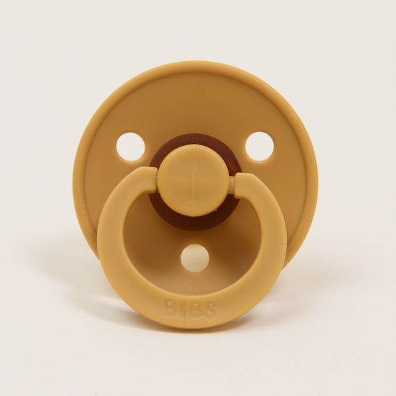 The Bibs Pacifier 2 Pack in Mustard features a classic design with a tan hue, round shield, and three holes. It includes a circular handle embossed with "BIBS," making it ideal for helping your little one self-soothe. The pacifiers are displayed upright against a plain white background.