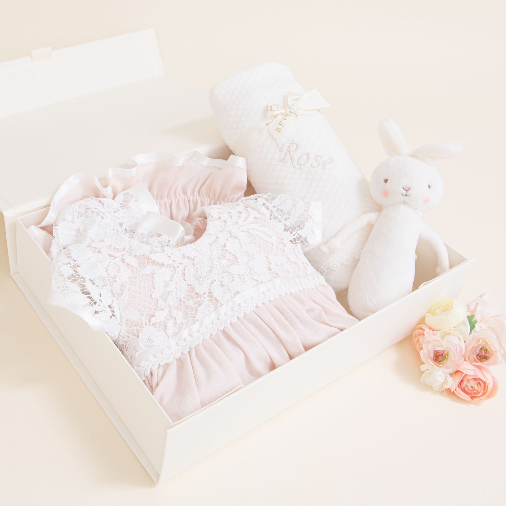 The Rose Newborn Gift Set - Save 10% contains a pink lace baby dress, a white baby blanket made of pima cotton with the name "Rose" embroidered, and a white stuffed bunny toy. Beside the box, there is a small bouquet of pink and white flowers.