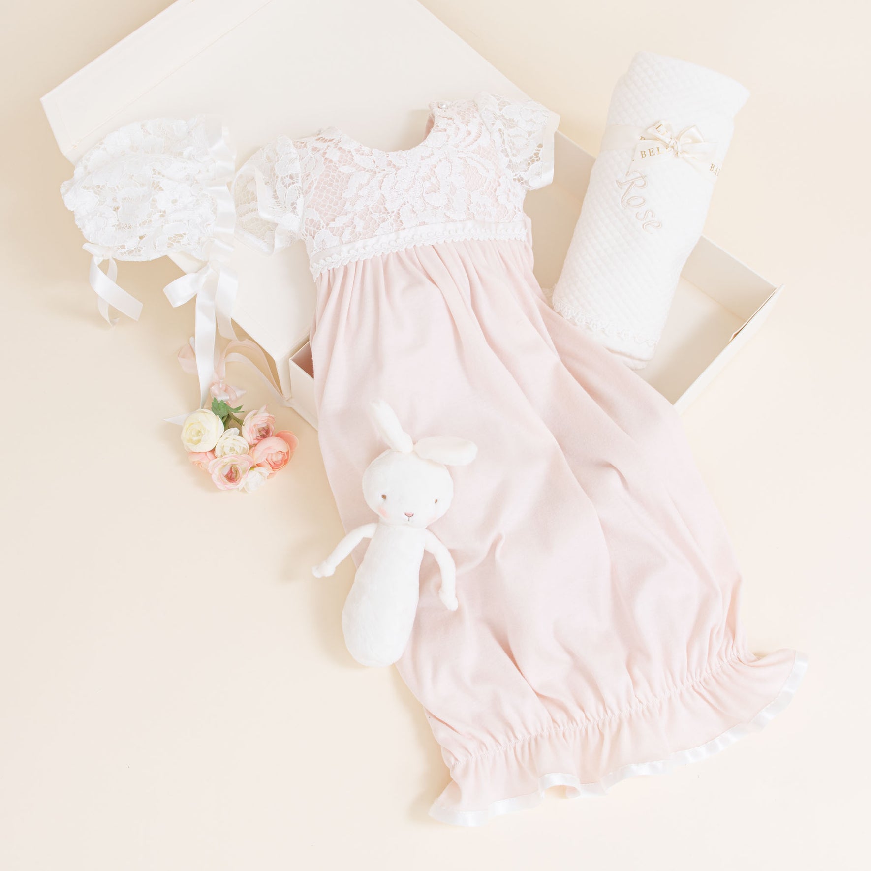 Rose Newborn Gift Set - Save 10% displayed in a box. Includes a pale pink newborn coming home gown with a rose patterned lace bodice, matching bonnet, white plush bunny toy, quilted blanket, and a small bunch of pastel-colored roses. The box is open on a light beige surface.