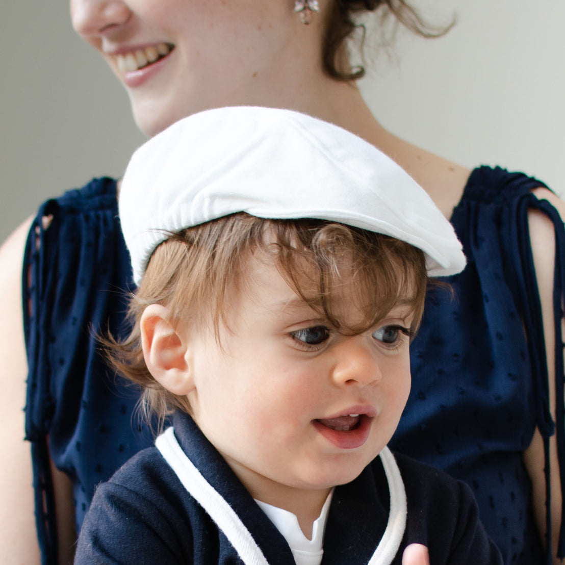 Closeup photo of a baby boy sitting with his mother and wearing the white Elliott Newsboy Cap and Elliott Jacket.