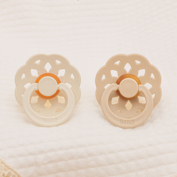 The Chloe Pacifier Set in Ivory & Vanilla includes two baby pacifiers. Each pacifier is set on a textured white surface, one showcasing a bohemian-inspired design with a cream and orange center, and the other crafted from natural rubber in pale beige with a tan center. This set is perfect for helping little ones self-soothe.