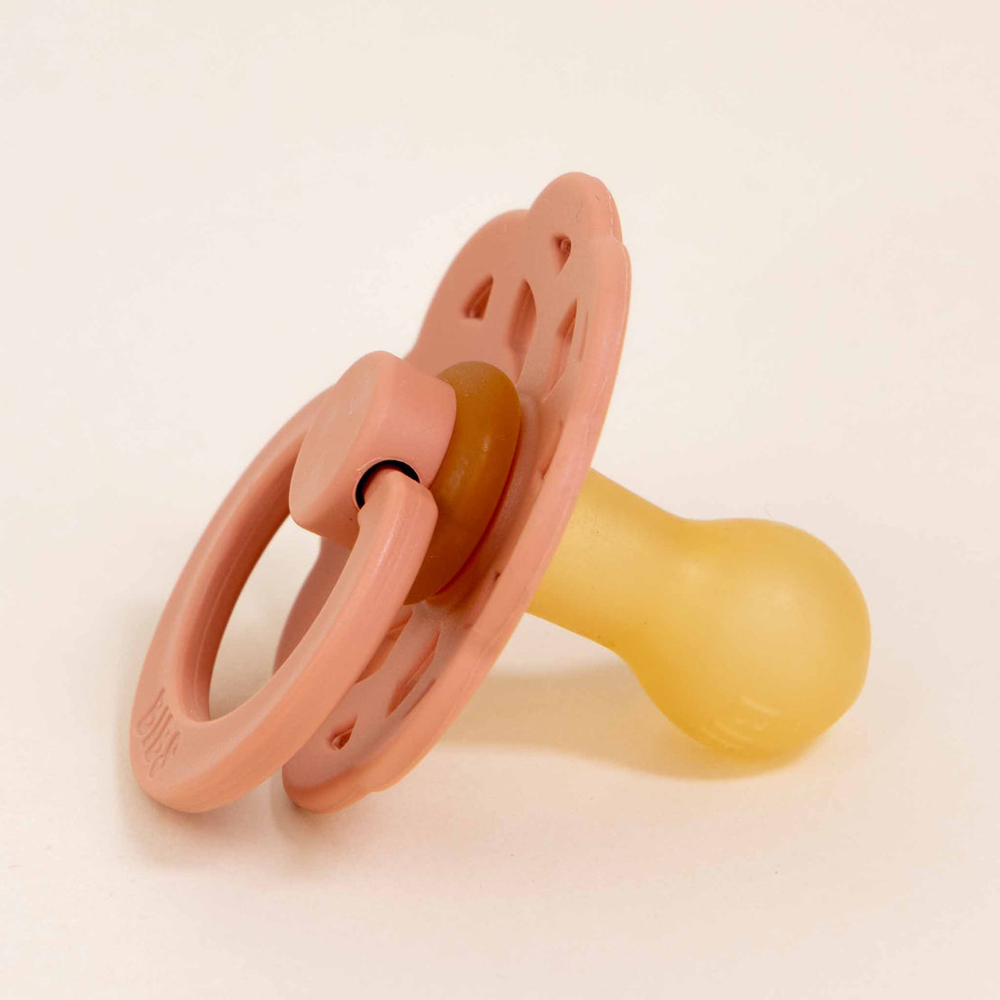 A peach Bibs Lace Pacifier, crafted from natural rubber and featuring a circular handle with a rounded nipple, is lying on a white surface. The pacifier exudes a touch of bohemian-inspired design, reminiscent of Denmark’s minimalistic style.