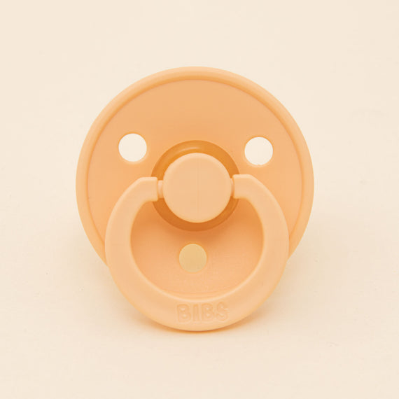 A Bibs Pacifier from the Peach Sunset 2 Pack, crafted from natural rubber, features a round shield with three ventilation holes and a ring handle embossed with the "BIBS" brand. This Danish-designed pacifier is ideal for helping little ones self-soothe, set against a light cream background.