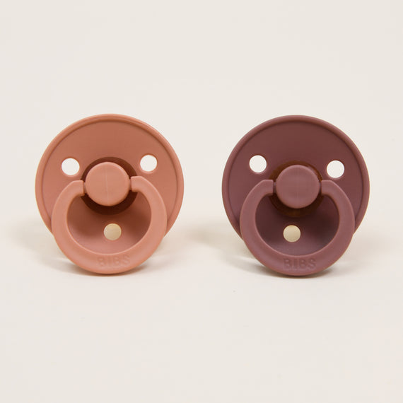 A pair of Bibs Pacifier Set | Peach Sunset & Woodchuck pacifiers is shown side by side against a plain background. The pacifier on the left features a light peach color, while the one on the right boasts a darker mauve shade. Both have circular shields with three holes and a ring handle, designed to help babies self-soothe.