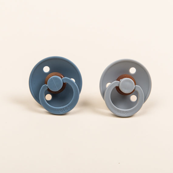 Displayed against a plain background are two baby pacifiers from the Bibs collection, available in Petrol and Cloud colors. These natural rubber pacifiers, manufactured in Denmark, are BPA-free and feature a round base with an embossed "BIBS" name. One pacifier boasts a darker blue shade than the other.