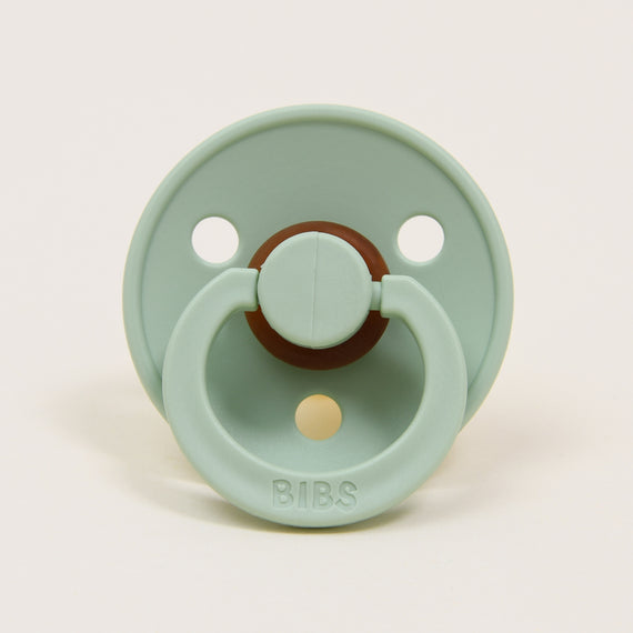 A pistachio-colored natural rubber baby pacifier with a round shield and an attached ring. The brand name "Bibs" is embossed on the bottom of the shield, which features two ventilation holes. Designed in Denmark, it's perfect for helping your baby self-soothe.