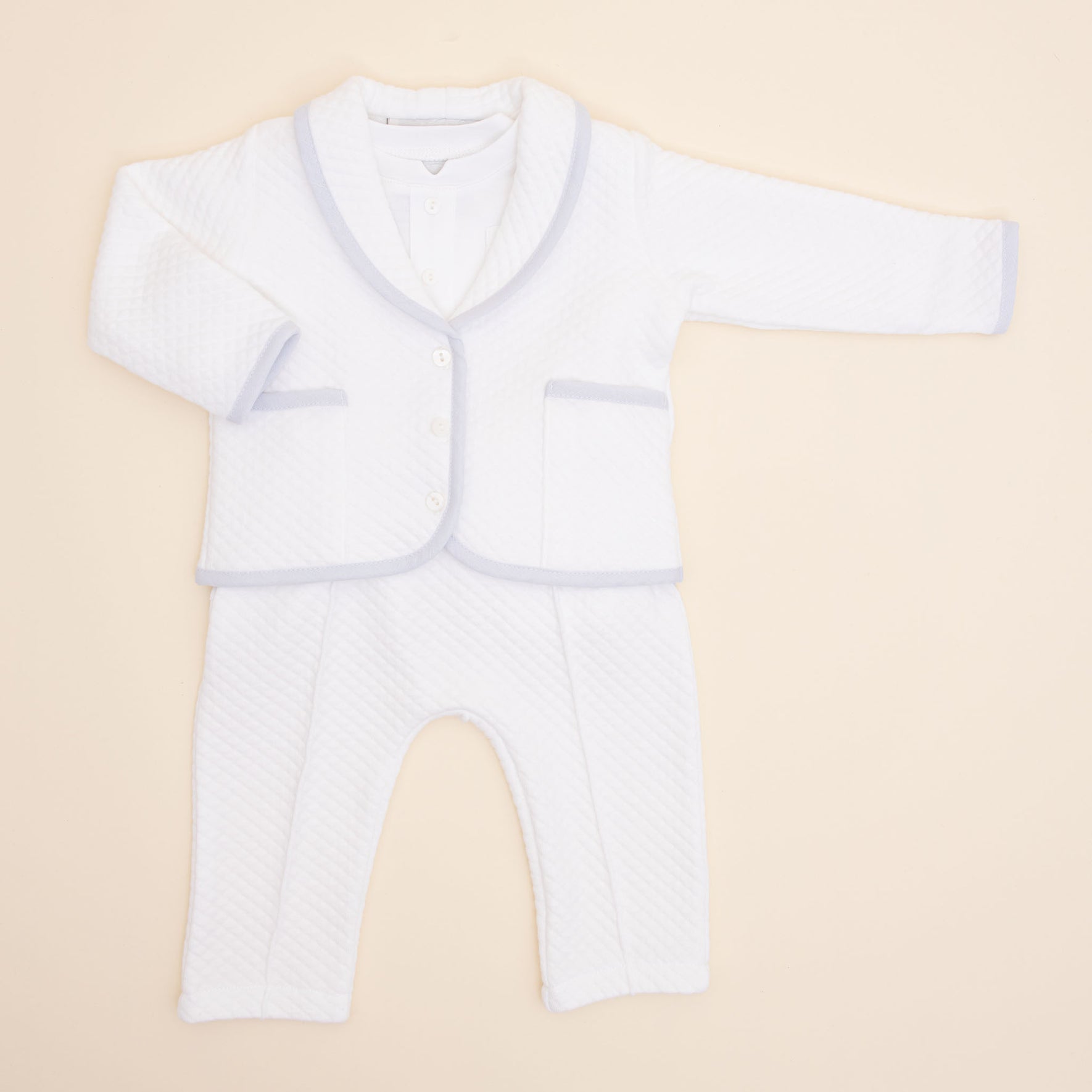 The Harrison Quilted Newborn Suit, an ideal choice for a baptism outfit, is displayed on a light beige background. This white ensemble features a long-sleeved jacket with buttons and matching pants. The quilted texture, complemented by light gray trim on the edges, adds an elegant touch suitable for special occasions.