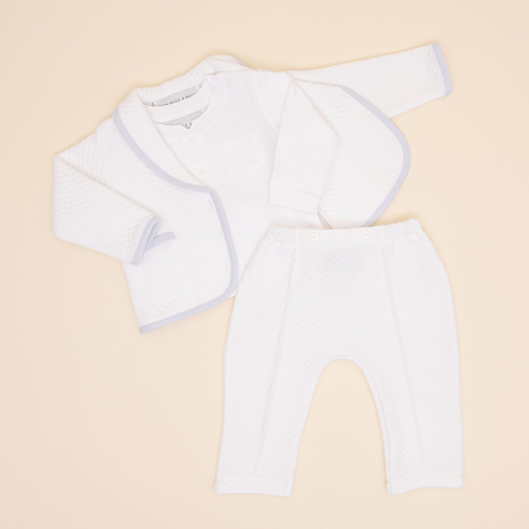 The Harrison Quilted Newborn Suit is beautifully displayed on a light surface, showcasing a quilted white jacket with subtle trim, a white top, and matching quilted pants. This adorable ensemble is perfect for newborn christening clothes, with each piece arranged to highlight its front.