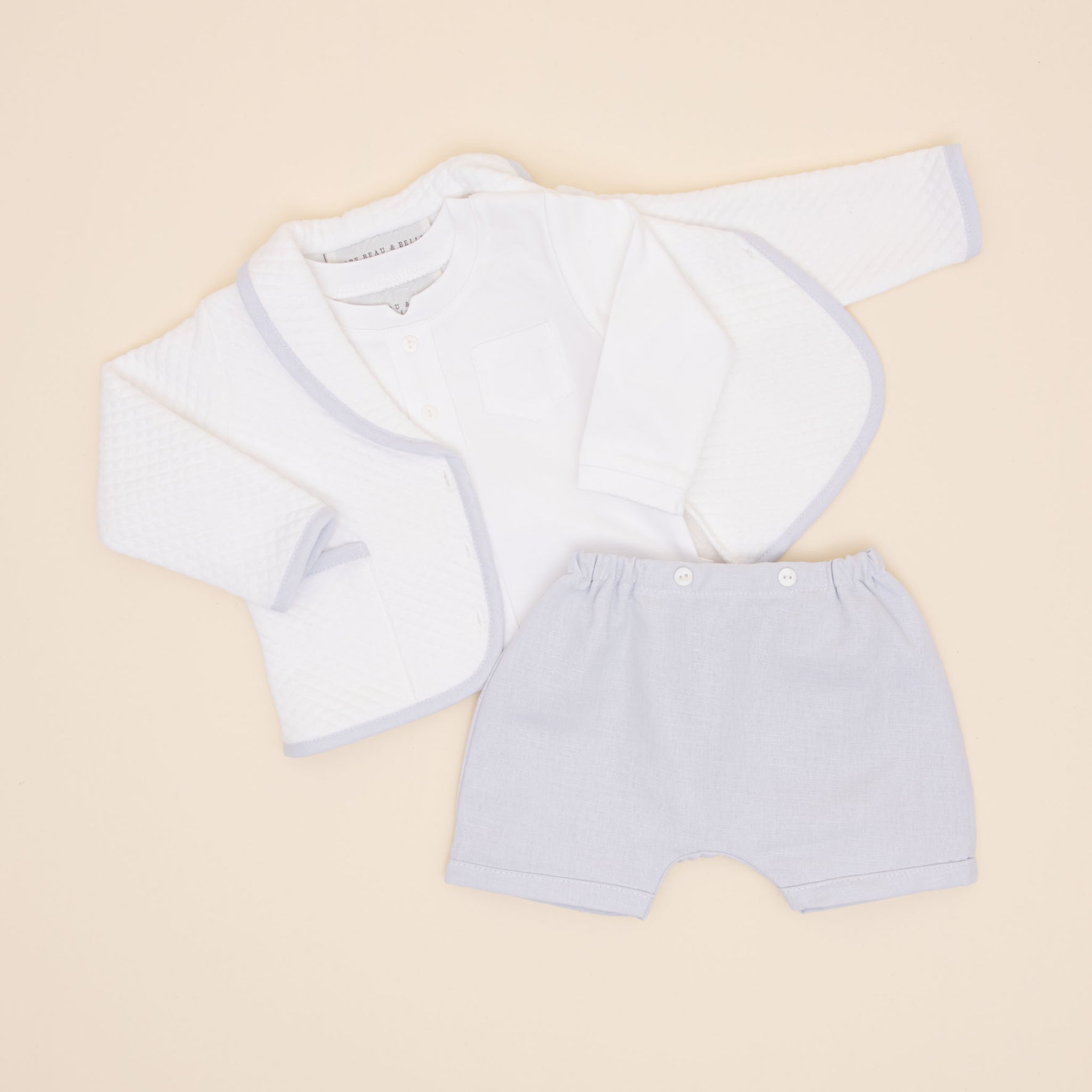 The Harrison Quilted Newborn Suit, a neatly arranged baptism outfit consisting of a white quilted jacket, a white long-sleeved shirt, and light gray shorts, is elegantly displayed on a light beige background.