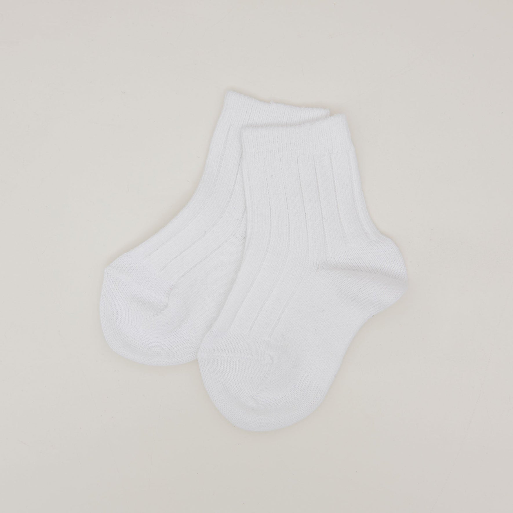 A pair of Ribbed Socks, crafted from a white cotton blend and made in Spain, is displayed flat against a simple light background. The socks have a soft appearance with distinct ribbed detailing, and the toes and heels are designed with a subtle rounded shape.