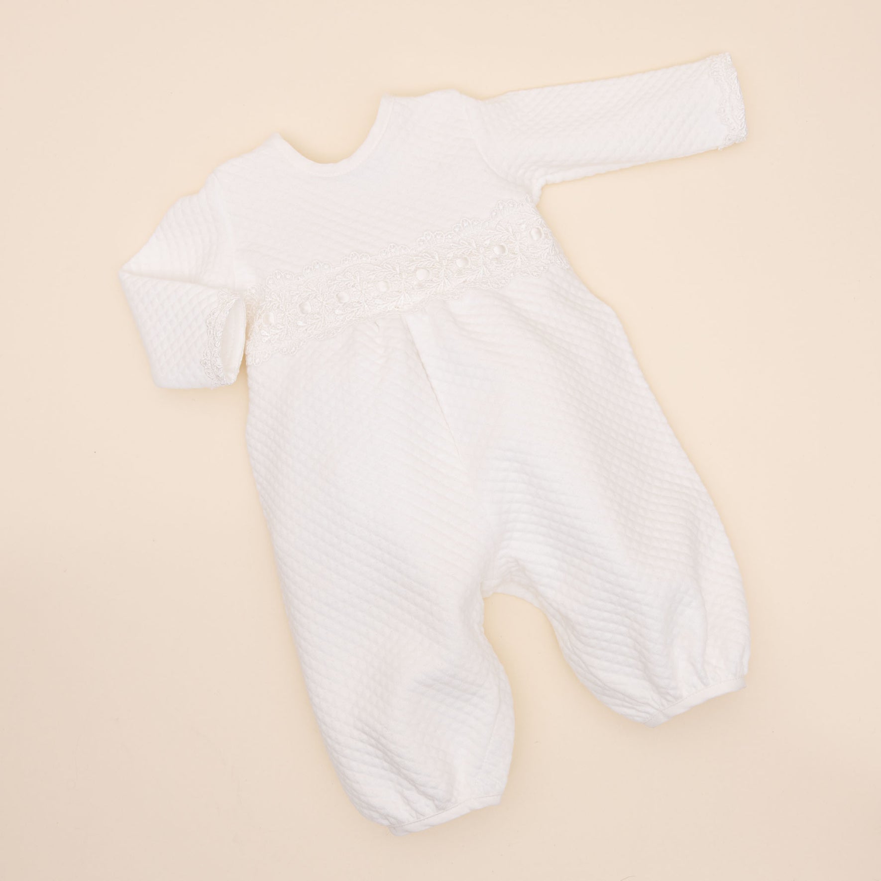 A white, long-sleeved Madeline Quilted Newborn Romper is laid out flat on a beige background. The onesie, handmade in the USA, has a textured pattern and features lace detailing across the chest area. The garment has a round neckline and closed feet.