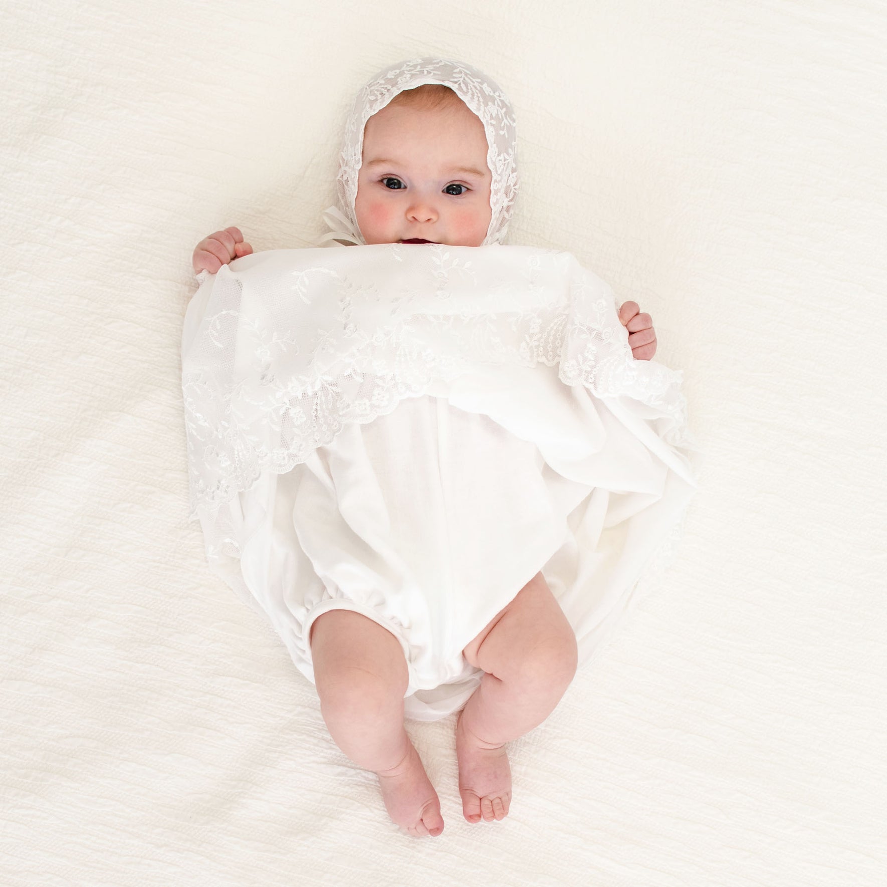 A baby girl holding up her skirt showcasing the detail under lace hem of the Ella lace baptism dress.