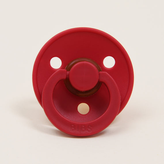 The Bibs Pacifier 2 Pack in Ruby features a classic design with a red circular shield that includes three holes and a natural rubber teat. The handle is attached to the front, and "BIBS" is embossed at the bottom, making it an ideal choice for helping babies self-soothe.