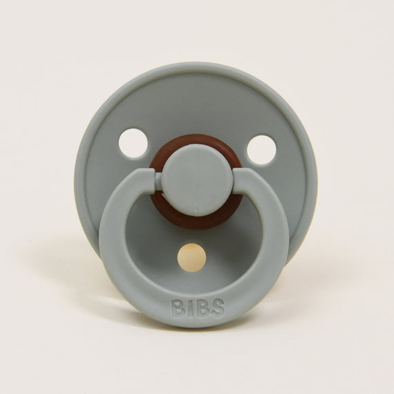 The Bibs Pacifier in Sage is elegantly designed with a gray tone and classic features, including two circular holes in the shield and a round, brown center crafted from natural rubber. The handle attached to the shield has "BIBS" embossed on it. Displayed upright against a white background, it is ideal for helping babies self-soothe.