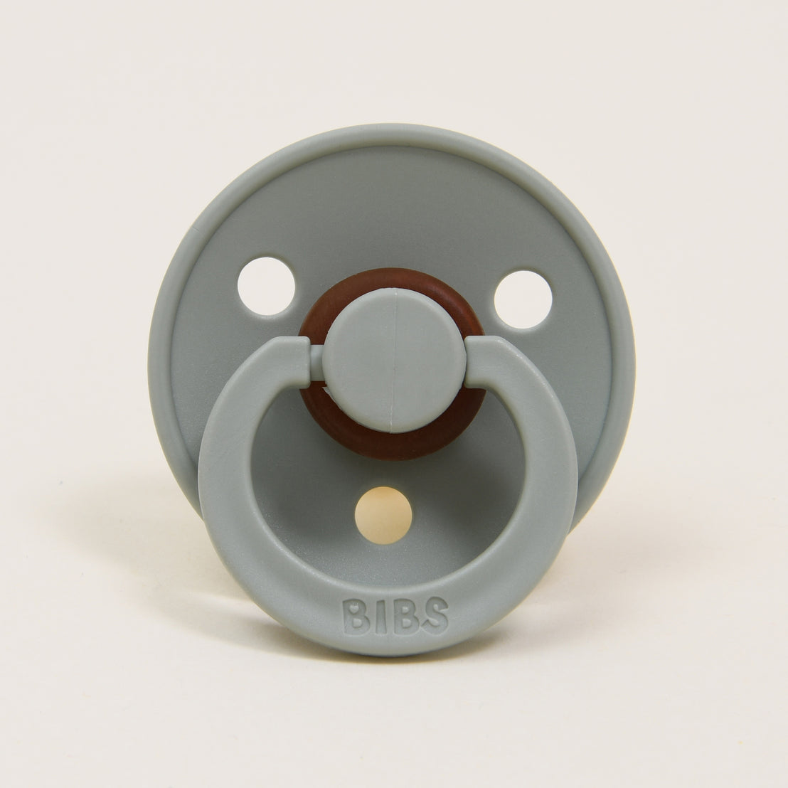 Set against a plain background, a timeless and elegant pacifier in the color gray is showcased, featuring three ventilation holes and a circular handle. Crafted from natural rubber, this BIBS pacifier has "BIBS" embossed on the front, providing style and comfort to help your baby self-soothe.