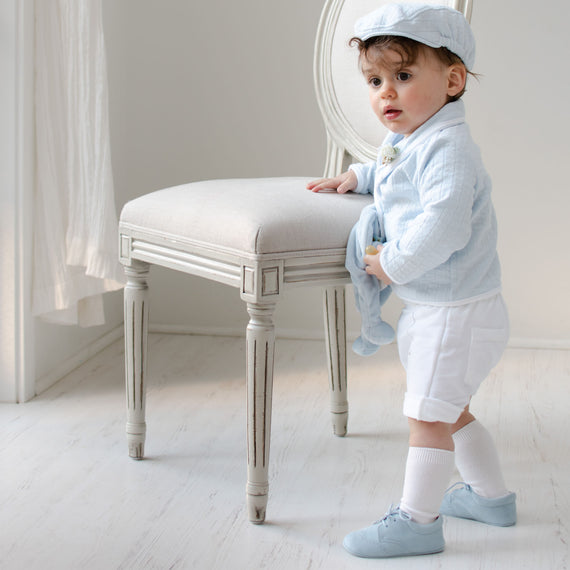 Baby boy standing by a chair and wearing the Logan 3-Piece Shorts Suit (with matching Newsboy Cap).