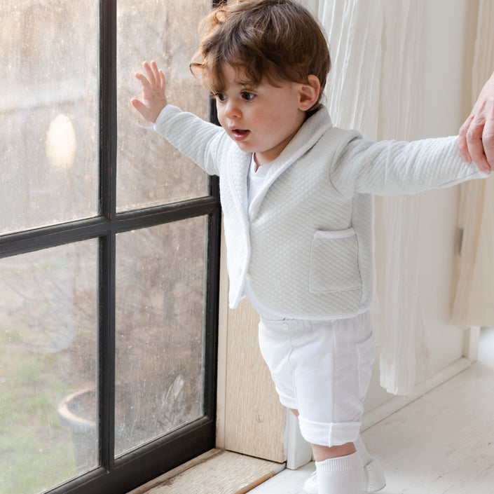 A young child stands by a window, dressed in a special occasion outfit featuring the Asher Soft Teal 3-Piece Shorts Suit. The child's hands are held by an unseen individual, and the room's white walls and curtains add to the tender scene.