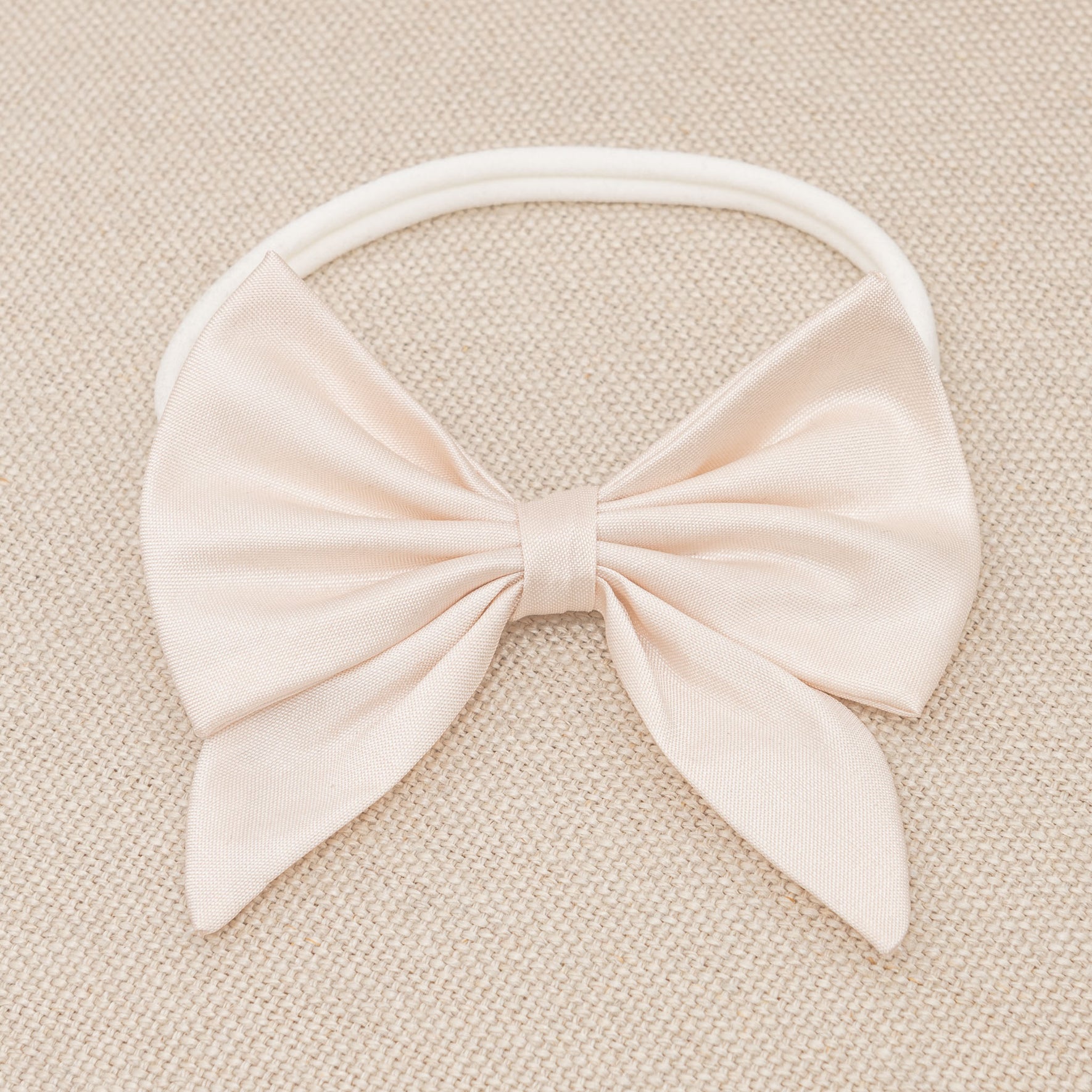 A pale pink fabric bow from the Elizabeth Accessory Bundle - Save 15%, perfect for a baptism dress, is attached to an elastic headband. The bow is neatly tied with two tails, and the headband lies on a textured beige surface.