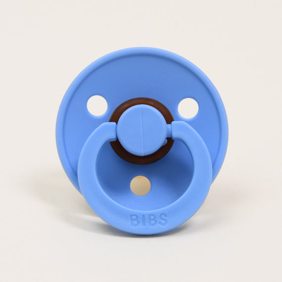 A sky blue pacifier with a round shield and a brown center ring, made from natural rubber to ensure comfort and safety. The handle is embossed with the "BIBS" brand name, and its classic and sophisticated design includes three circular holes against a plain white background.