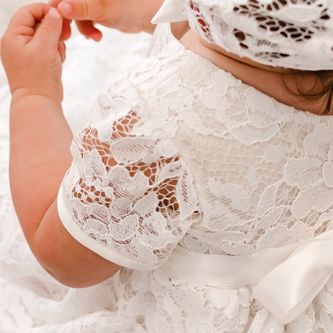 Close-up of a baby wearing a Rose Christening Gown & Bonnet, with a focus on the delicate fabric and a small hand reaching out gently.