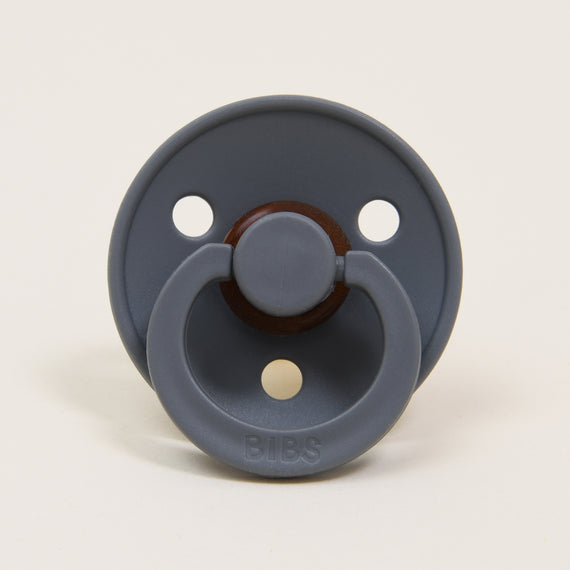 The Bibs Pacifier 2 Pack in Smoke features a natural rubber design made in Denmark, showcasing a dark gray round shield with three small holes and a central circular knob. The brand name "BIBS" is elegantly embossed on its lower part, ensuring a quality design that helps your little one self-soothe.