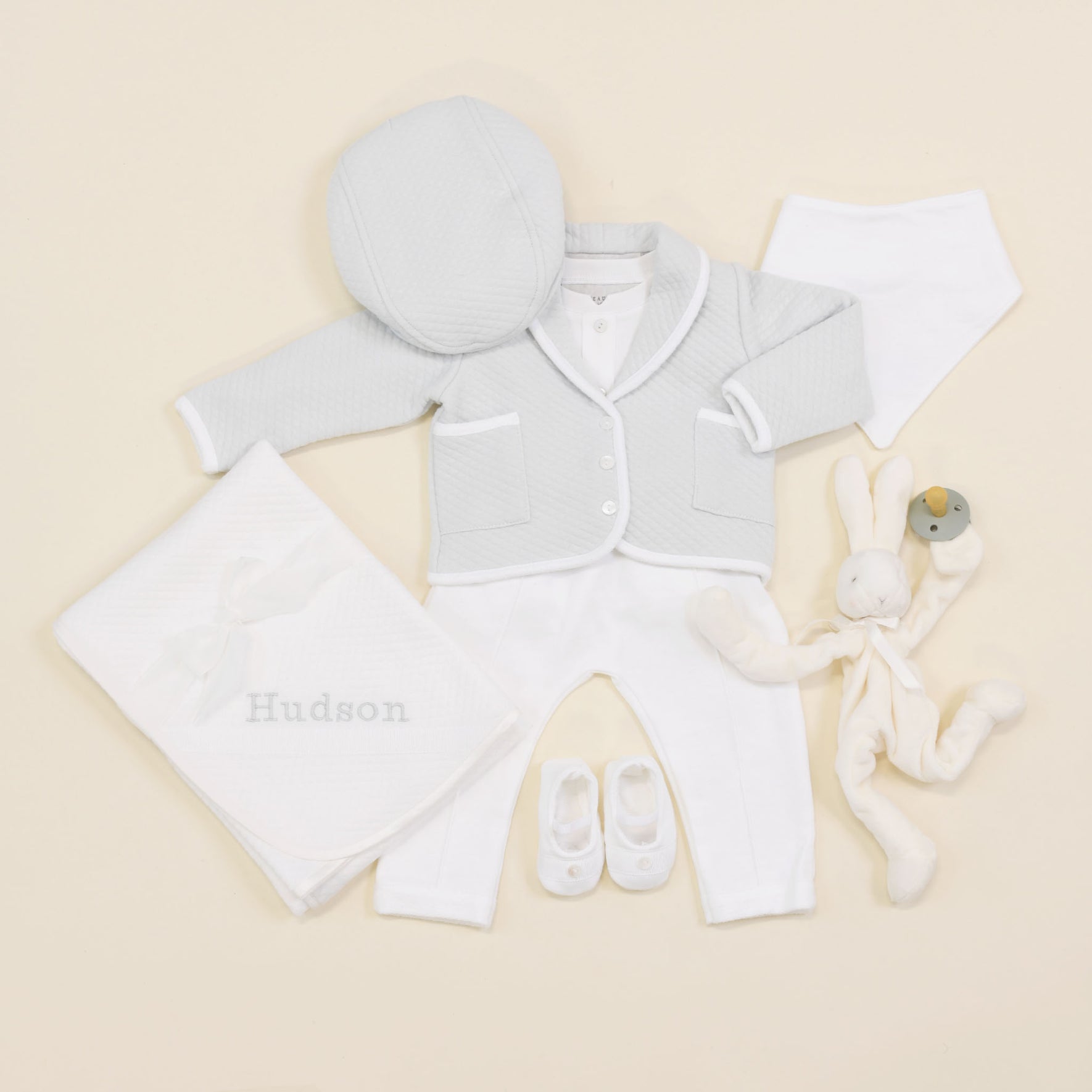 The Asher Soft Teal 3-Piece Suit includes a cozy light gray quilted cotton jacket, a matching hat, white pants, and small booties. Additionally, it comes with a soft white blanket featuring "Hudson" embroidery and a stuffed rabbit toy with a rattle.