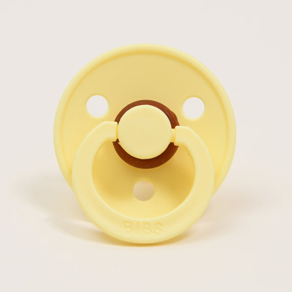 The "Bibs Pacifier 2 Pack | Sunshine" set includes a yellow pacifier made from natural rubber, featuring a classic design with a round shield and two holes on a plain white background. The central part and the ring are slightly darker in color, with the "BIBS" brand name embossed on the ring handle, ensuring comfort for babies as they self-soothe.